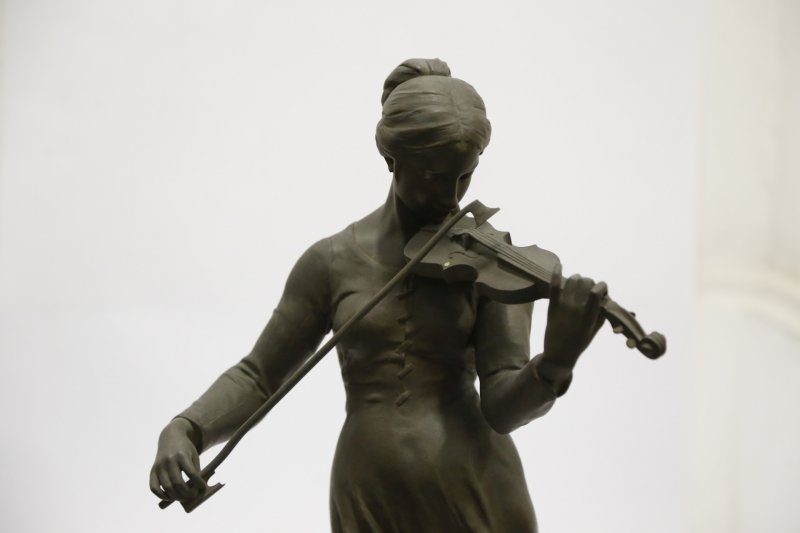 Fotografia de uma escultura em bronze de uma mulher tocando violino, intitulada 'A Violinista', criada pelo escultor Alfred Boucher. A figura apresenta detalhes realistas, com expressão serena e concentração no ato de tocar. Ela segura o arco com a mão direita enquanto mantém o violino apoiado no ombro esquerdo. A mulher veste um traje simples e seu cabelo está preso para trás. A escultura transmite um momento de serenidade e dedicação à música.