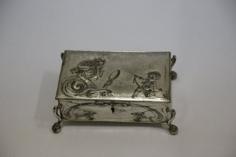 A imagem mostra uma caixa decorativa em metal com detalhes em relevo, em estilo art noveau. Na tampa da caixa, há uma figura de uma mulher de cabelos longos segurando um espelho, acompanhada por um cupido que segura um colar de pérolas. A caixa possui pés ornamentados que se assemelham a patas de animais. Possui decoração com fitas e flores arrematando a fechadura, sem chave.