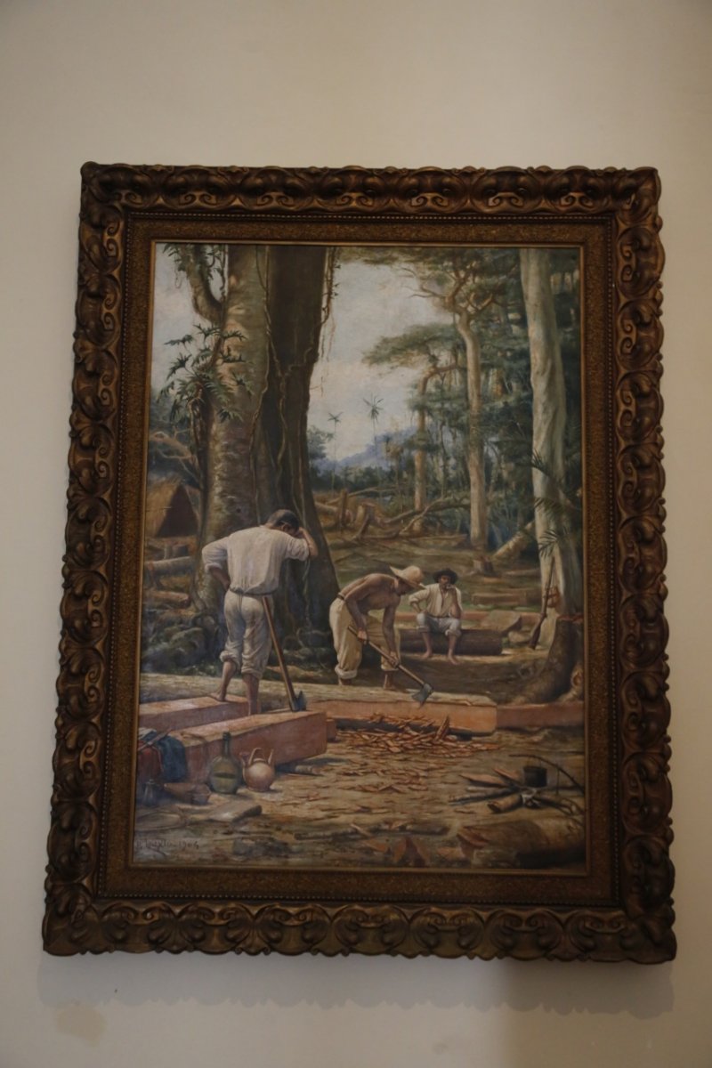 A pintura retrata uma cena de trabalho manual em uma floresta. Três homens estão envolvidos em atividades de corte de madeira. Dois deles estão de pé, um segurando um machado e o outro uma ferramenta de corte, enquanto um terceiro homem está sentado em um tronco ao fundo, fumando. No chão, há lascas de madeira espalhadas e alguns objetos, como garrafas e uma panela. A floresta ao fundo é densa, com árvores altas e vegetação abundante. A tela possui uma moldura ornamentada com padrão fitomórfico