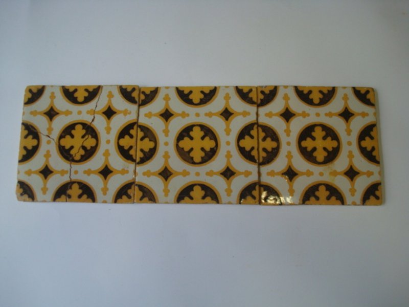 A imagem mostra três azulejos decorados dispostos em linha, formando um padrão contínuo. Os azulejos têm um fundo branco com desenhos circulares em amarelo e marrom. No centro de cada círculo há uma forma que lembra uma cruz ou flor estilizada em amarelo, cercada por um anel marrom. Entre os círculos, há pequenos losangos amarelos formando um padrão repetitivo e simétrico