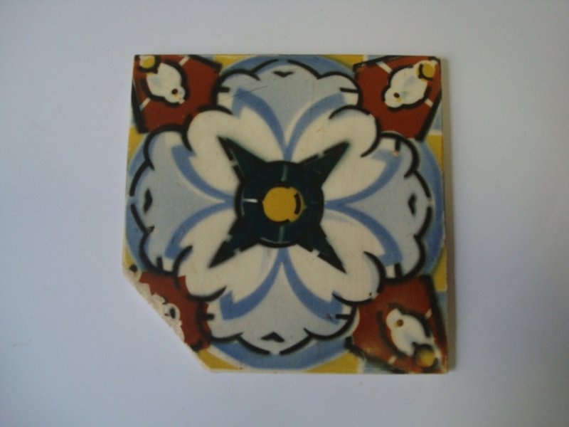 Fotografia de um azulejo em faiança com elementos decorativos em padronagem floral estilizada pintada a mão. O centro do azulejo possui um desenho simétrico de uma flor com pétalas brancas e detalhes em azul claro, delineadas em preto. No centro da flor, há um círculo amarelo cercado por um desenho em preto, que lembra uma estrela. Nos cantos do azulejo, há formas em vermelho e branco, também delineadas em preto, que parecem ser flores ou elementos decorativos complementares. O azulejo apresenta uma pequena lasca no canto inferior esquerdo, revelando sinais de desgaste